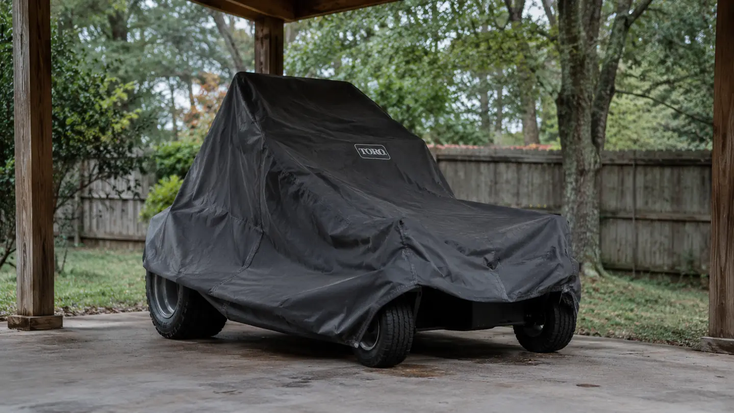 A zero turn mower parked under a carport with a cover on, trees in the background