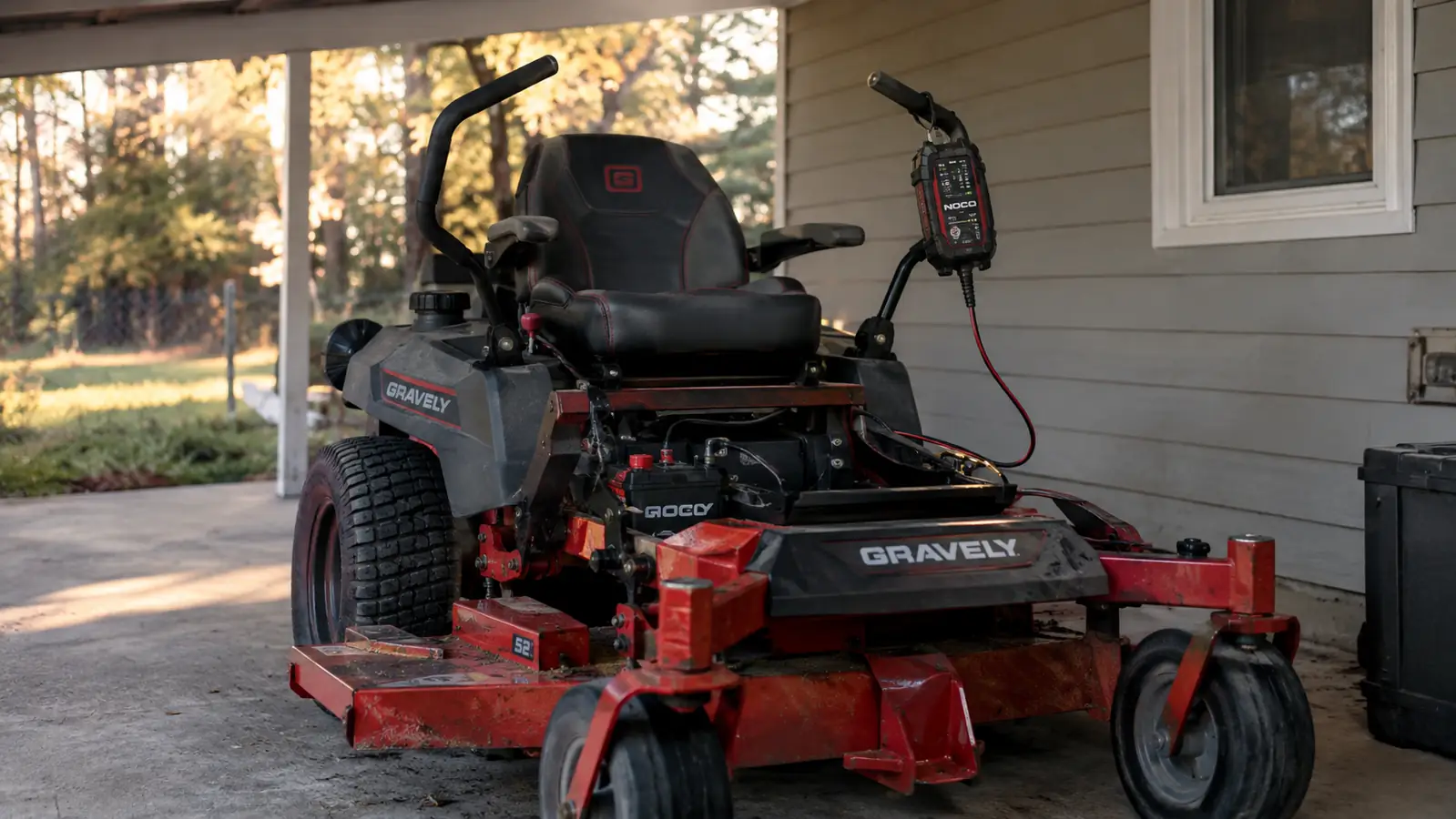 A zero turn mower parked under a carport with a maintainer cord running to the battery