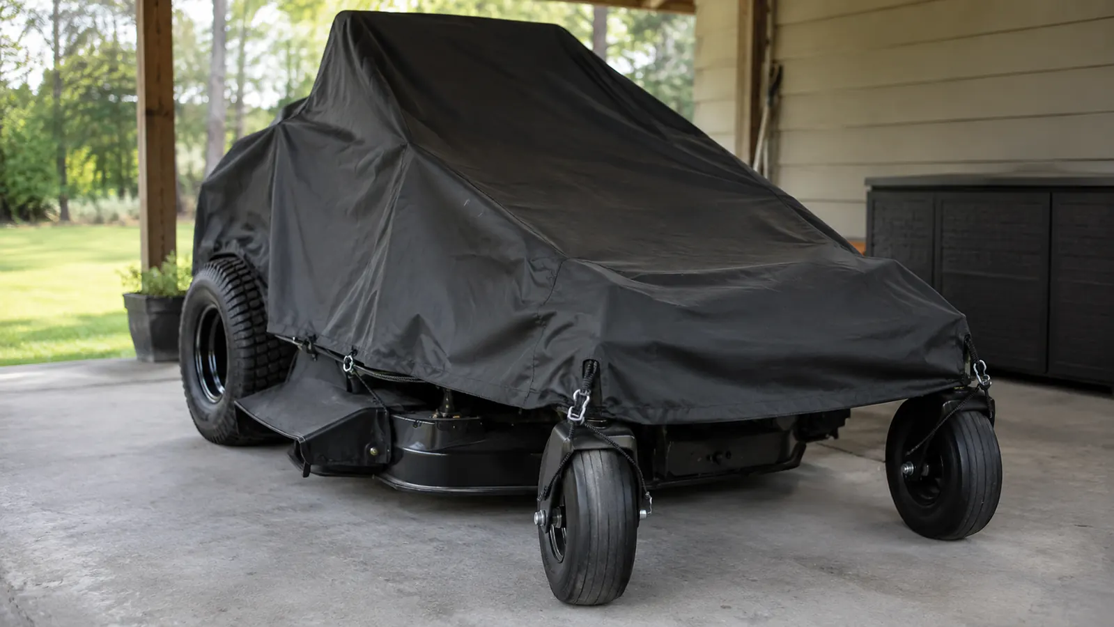 A zero turn mower parked under a carport with a cover secured by a bungee cord at each corner